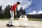 image of golf  - Young male golf player in red shirt and grey pants chipping golf ball out of a sand trap with sand wedge and sand caught in motion - JPG 