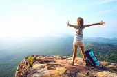 stock photo of woman  - Young happy woman with backpack standing on a rock with raised hands and looking to a valley below - JPG 