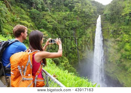 Picture or Photo of Couple tourists on Hawaii by waterfall. Tourist taking photo pictures of Akaka Falls waterfall on Hawaii, Big Island, USA. Travel tourism concept with multicultural tourist couple.