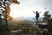 stock photo of valley  - Young hiker with backpack standing with raised hands on a cliff - JPG 