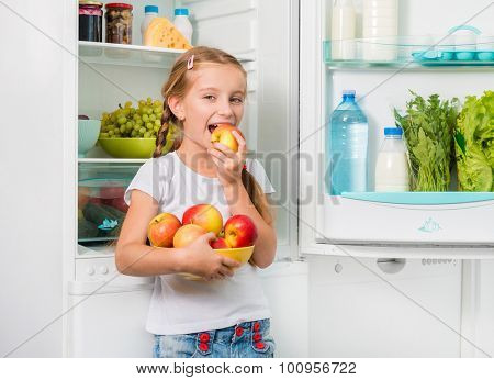 little girl biting an apple near opened fridge