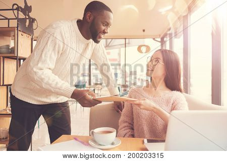 Open it. Cheerful afro american student giving a letter to her beautiful positive friend while sitting in the cafeteria