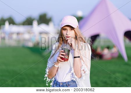 Stylish young girl stands with a cocktail in her hand against the backdrop of a park. Summer concept. Rest in the park