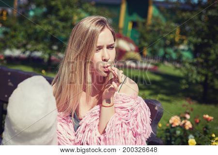 Instagram filter. Close up portrait of tender blond student eating white huge candy cotton with passion. Young & carefree beautiful female hipster traveler is taking rest outdoors in summer city park.