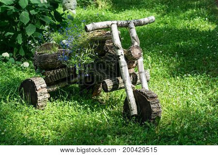 In the forest in the clearing stands a motorcycle from wooden logs. A motorcycle with a sidecar or bassinet. The carrycot and seat are baskets with flowers.