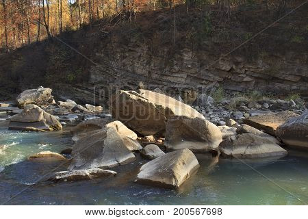 Mountain creek with turquoise water and big stones in it in front of a layerd rock with orange forest