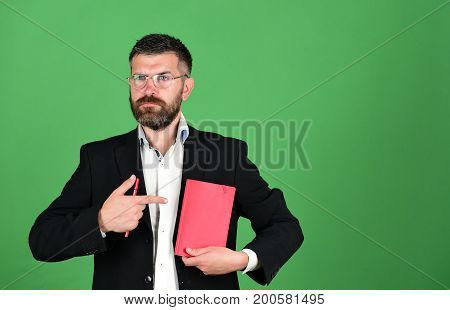 Knowledge and schedule concept. Professor with serious face expression. Notebook in red color in guys hands on green background. Teacher wears glasses and points at organizer. Man with beard and book