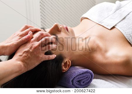 Head and face massage in spa. Female beautician work in wellness center. Professional massagist make relax healthy procedure to beautiful indian girl in cosmetology cabinet or beauty parlor.