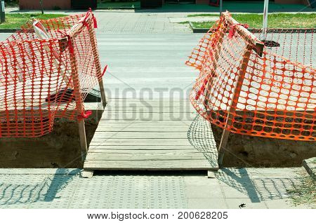 Improvised wooden bridge over the trench on the street reconstruction site for pedestrians and orange construction safety fence net.