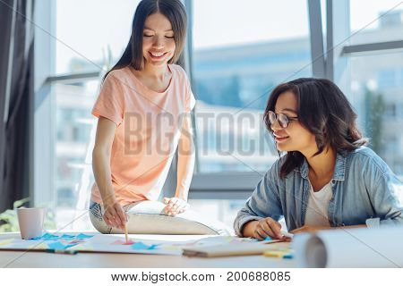Informal atmosphere. Joyful delighted positive woman sitting on the table and holding a pencil while explaining her plan to a friend