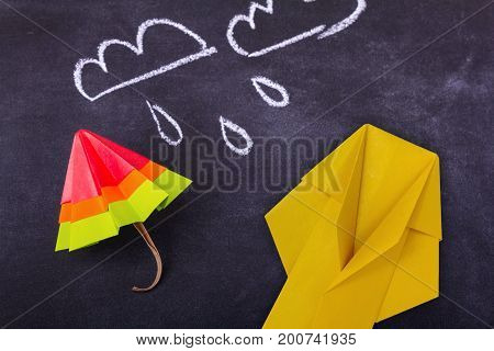 Origami cartoon protection umbrella and raincoat on a blackboard background with drops and clouds