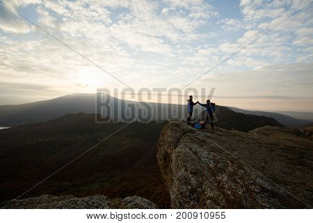 Two climbers standing on summit above clouds in the mountains holding hands. Silhouettes of hikers celebrating ascent on sunset. Cooperation, synergy, friendship concept.