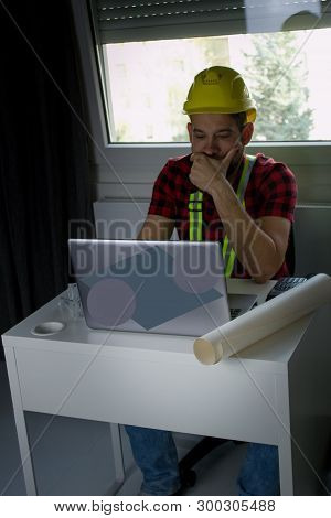 Bored Construction Worker Is Sitting At His Desk At The Computer, Looking At Building Plans And Coun