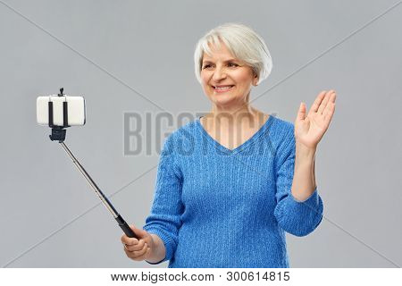 technology and old people concept - smiling senior woman taking picture by smartphone on selfie stick waving hand over grey background