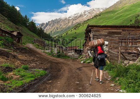 Tourist With Backpack And Trekking Poles Goes Up Rural Road Along Village To Mountains. Gorge Tersko