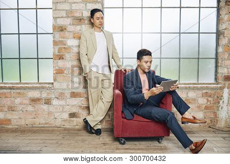 Young Handsome Entrepreneur Reading Article On Tablet Computer And His Coworker Looking Over His Sho