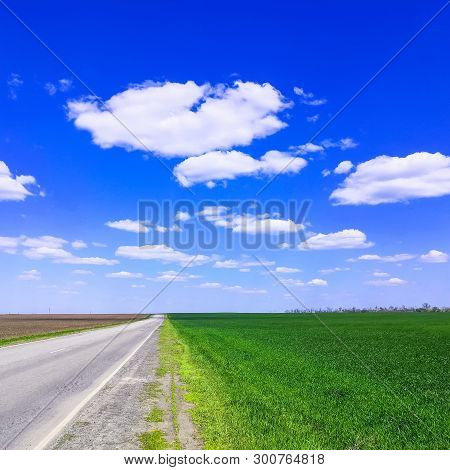 Empty Highway Through Green Grass Field Under White Clouds On Bright Blue Sky In Spring Summer Day. 