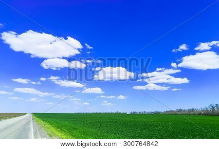 Empty Highway Through Green Grass Field Under White Clouds On Bright Blue Sky In Spring Summer Day. 