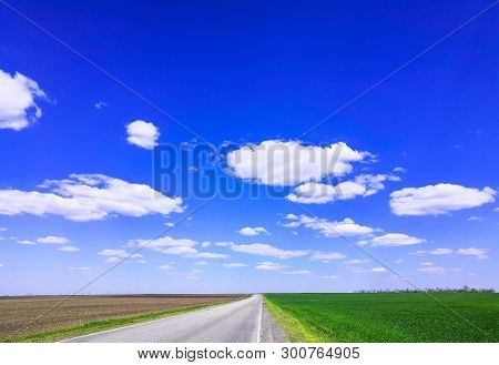 Empty Highway Through Green Grass Field Under White Clouds On Bright Blue Sky In Spring Summer Day. 