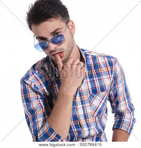 Sensual young man romantically looking at the camera and touching his lips while wearing a pair of sungalsses and a checkered shirt on white studio background