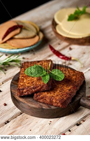 closeup of some cooked tofu fillets on a round wooden tray placed on a white rustic table, next to some plates with slices of vegan cheese, and some cold cuts made of tofu