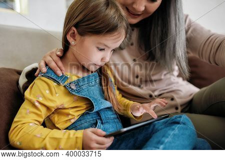 Little Caucasian Girl With Digital Tablet Sitting On Sofa At Home Next To Woman Babysitter, Cropped.