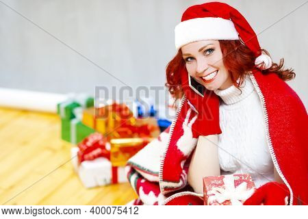 Gleeful Caucasian Santa Girl With Wrapped Present In Hand While Happily Talking By Cellphone And Sit