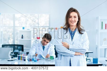 A Beautiful Young Scientist Stands Smiling In A Science Lab With A Male Scientist Sitting In The Bac