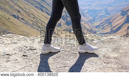 Woman In Black Jeans And Light Grey Sneakers Walks Along Rocky Cliff Edge Against Distant Mountains 