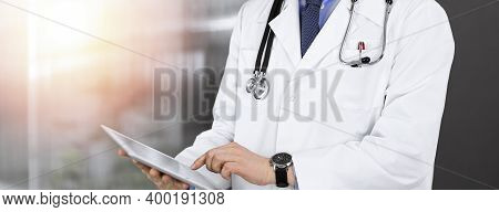 Unknown Male Doctor Iusing Tablet Computer In His Hands, While Standing In Sunny Clinic. Medicine Co