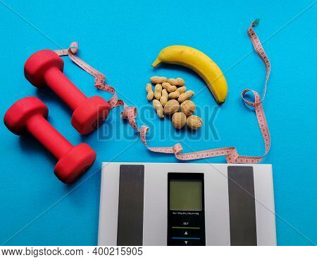 Electronic Scale, Banana, Nuts, Measuring Tape And Dumbbells On A Blue Background. Food And Fitness 