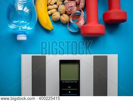 Electronic Scale, Bottle Water, Banana, Nuts, Measuring Tape And Red Dumbbells On A Blue Background.