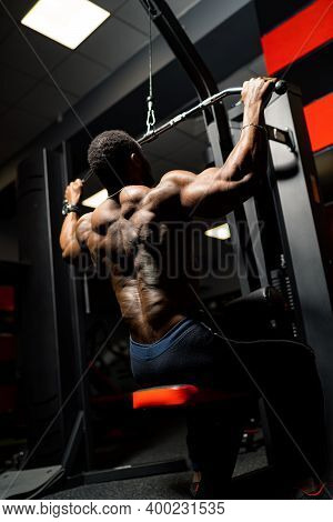 Athlete Pumps Biceps On The Simulator. African Handsome Bodybuilder In Gym. View From The Back.