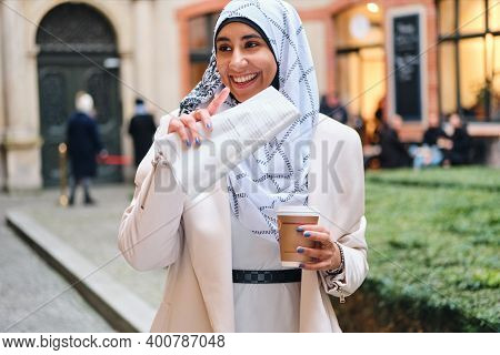 Young Cheerful Arabic Woman In Hijab Joyfully Walking Through Street With Coffee To Go And Newspaper