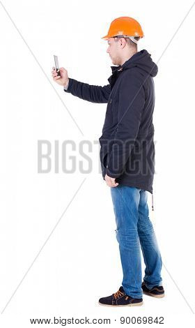 Backview of an engineer in construction helmet stands and using a mobile phone. Standing young guy.  Isolated over white background. Foreman photographs on the phone what he sees.