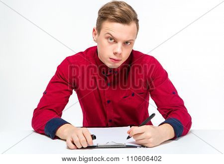Suspicious curious attractive young male in red shirt with raised eyebrow writing on clipboard over white background