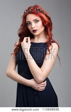 Beautiful red-haired fashion model posing in evening dress and in the diadem over dark background. f