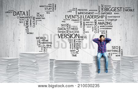Young man in casual wear sitting on pile of documents with business-related terms in form of world map on background. Mixed media.