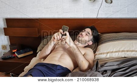 Young attractive man typing on the phone while lying on the bed at home in the morning when he just woke up