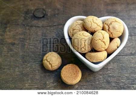 Italian Christmas cookies amaretti in a white heart shaped bowl.Tasty Italian amaretti biscuits on old wooden table.Amarettini cookies.Selective focus.
