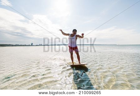 skimboarding, water sport and people concept - happy young man riding on skimboard on summer beach