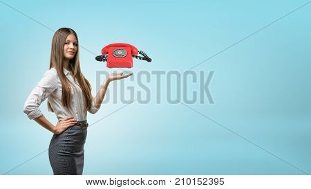 A blond businesswoman stands and holds one hand palm up with a red retro phone hovering above it. Telecom sales. Cold calls. Emergency and support number.