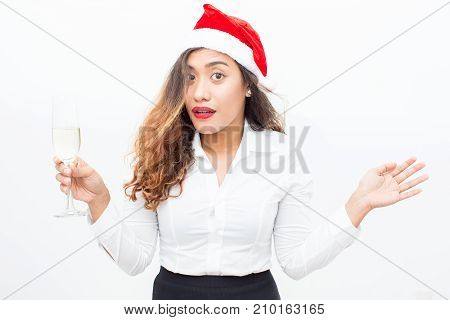 Portrait of confused young Asian businesswoman wearing Santa hat holding champagne flute and shrugging shoulders. Christmas and office party concept