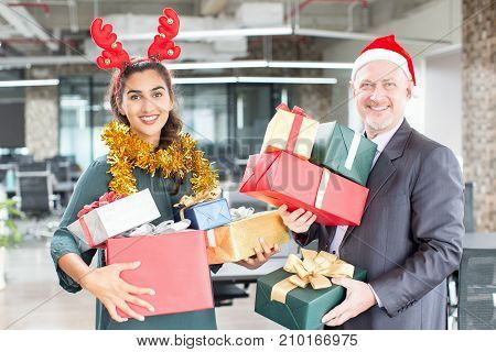 Joyful friendly colleagues enjoying Christmas time in office. Smiling happy business people with heap of Christmas presents looking at camera and ready for gift-giving. Holiday concept