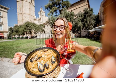 Young woman in red dress making selfie photo with sea Paella, traditional Valencian rice dish, sitting outdoors at the restaurant in the centre of the old town of Valencia