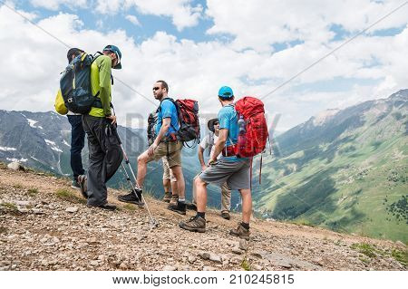 A group of hiker tourists with backpacks and tracking sticks rest and stands in the mountains listening to their guide against the background of green and snow-capped rocky mountains.