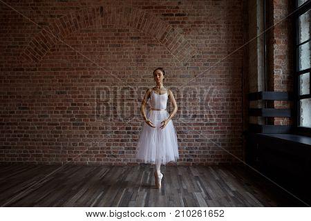 Attractive hardworking young ballerina wearing white pack and pointe shoes practicing center worrk standing on toes against red brick wall background with copy space for your advertising content