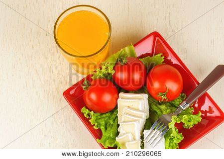 Tomatoes And Cheese Slices Lie On A Sheet Of Fresh Salad In A Red Plate. A Glass Of Orange Juice Sta