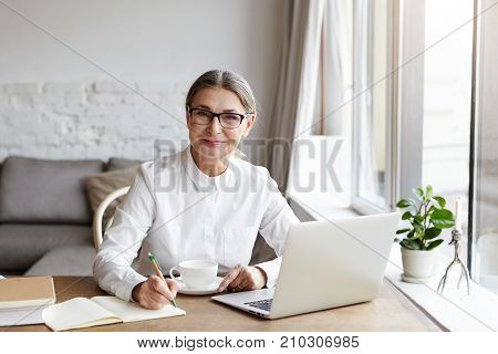 Attractive positive talented mature woman writer sitting in front of laptop and writing her new book about love looking at camera with joyfull smile. Senior female teacher making notes in copybook