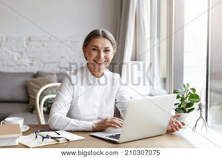 Happy mature woman psychologist or medical expert wearing white shirt working distantly on portable computer consulting her clients online. Senior people modern technologies job and occupation